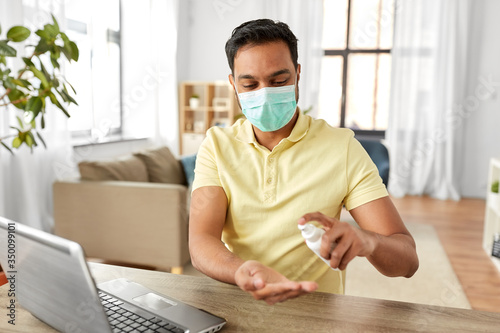 quarantine, remote job and pandemic concept - happy indian man wearing face protective medical mask for protection from virus disease with laptop computer using hand sanitizer at home office photo