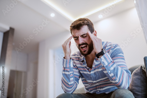 Young handsome bearded man sitting on sofa in living room, holding head and having headache.