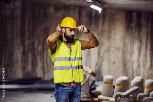 Photography Young bearded worker in vest putting his helmet on head while standing inside building in construction process