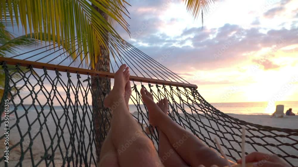 Slow motion of couple lying with coconut on hammock at beach, tourists relaxing against sky during sunset - Montego Bay, Jamaica