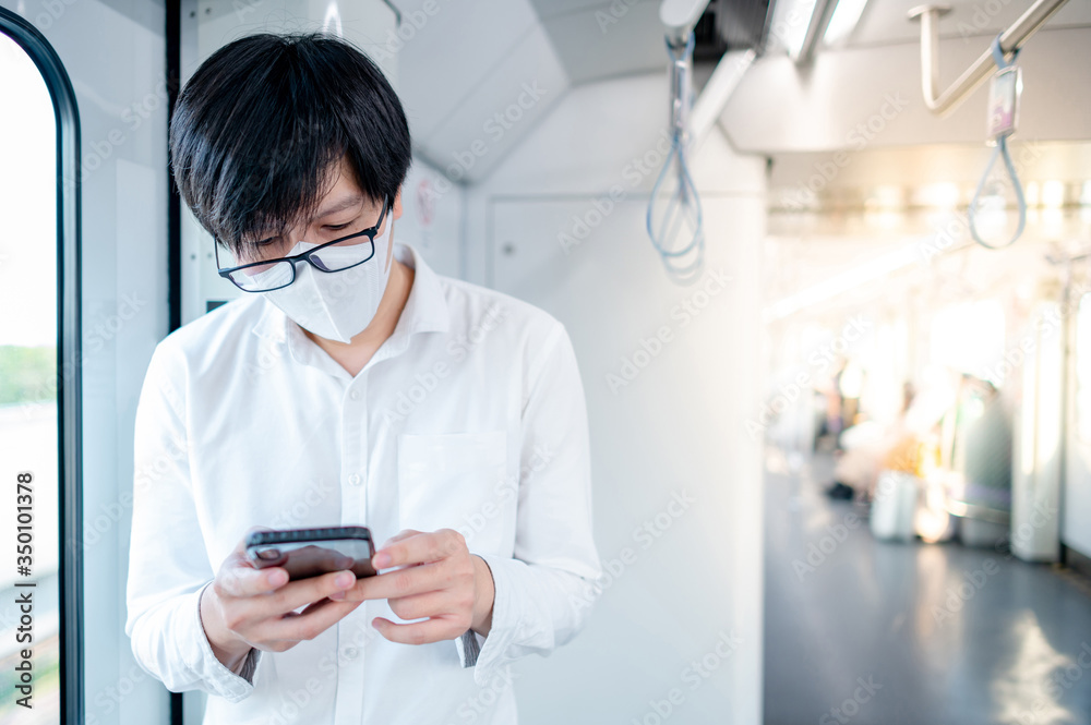 Asian man wearing surgical face mask using smartphone on skytrain or urban train. Coronavirus (COVID-19) outbreak prevention in public transportation. Social distancing for pandemic protection Asian man wearing surgical face mask using smartphone on skytrain or urban train. Coronavirus (COVID-19) outbreak prevention in public transportation. Social distancing for pandemic protection