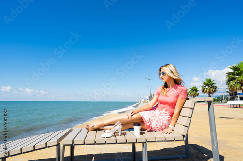 Young beautiful woman in red dress sitting on beach chair with shoes, coffee and looking on blue sea and sky.