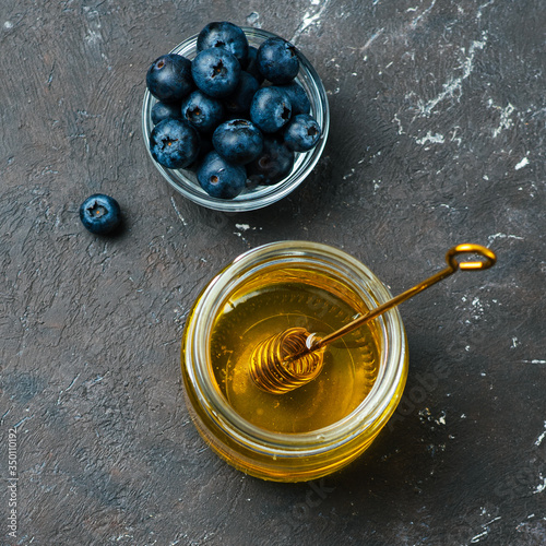 Healthy food ingredients - blueberries and honey. Glass jar with honey and dipper, fresh blueberries in small bowl over dark background. Top view or flat lay. Square crop. Copy space photo