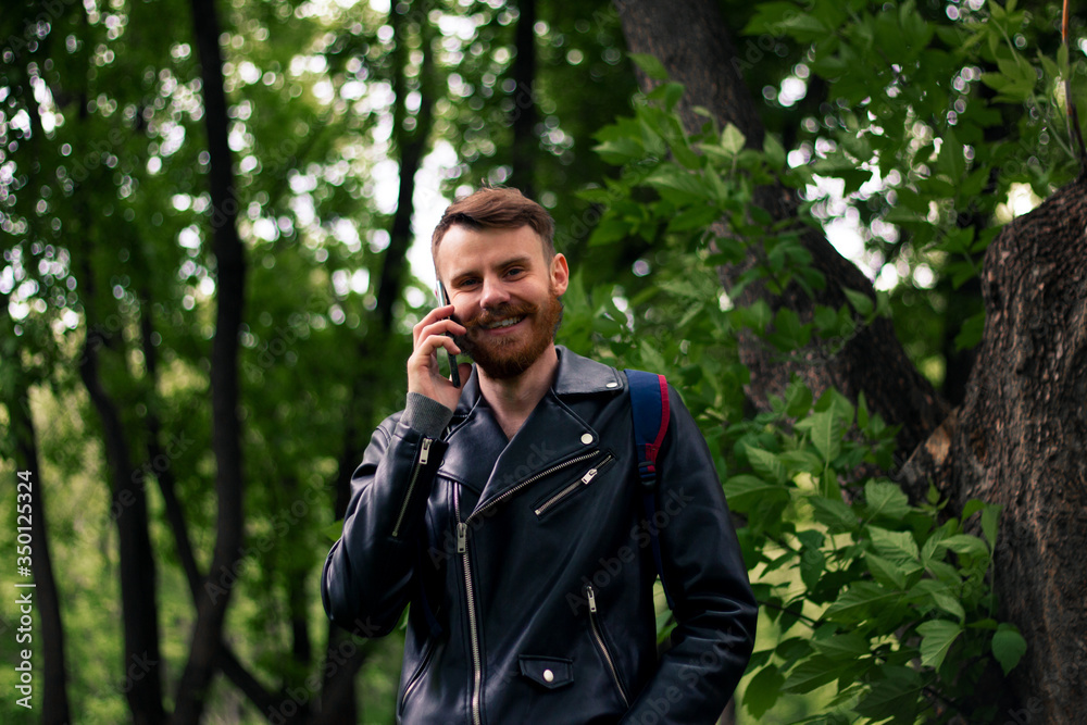 A man with a red beard smiles widely while talking on a smartphone while standing in a green forest in a black leather jacket. A man with a red beard smiles widely while talking on a smartphone while standing in a green forest in a black leather jacket.