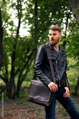 A young Irish guy with a brown bag over his shoulder and a thick beard looks into the distance while standing in the middle of the forest.