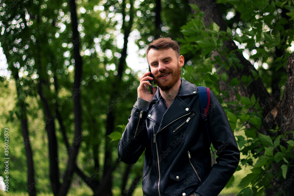 Smiling guy talking on a smartphone while standing in a forest in a leather jacket. Smiling guy talking on a smartphone while standing in a forest in a leather jacket.