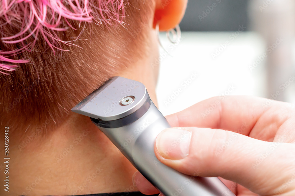Fototapeta premium Back view hairdresser hand is shaving pink hair of woman with electric trimmer in hair salon, close up.
