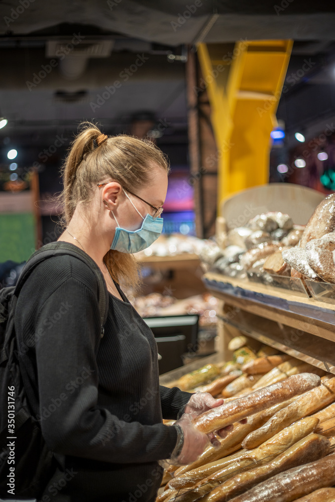 A young woman in a blue medical mask and transparent gloves buys bread in a supermarket. A woman is wearing a black sweater. Backpack. Woman with glasses. Covid-19.