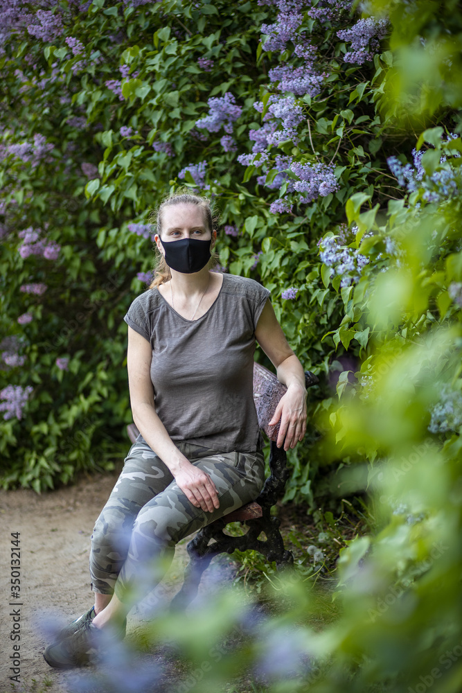 Portrait of a beautiful young woman in a black mask on a background of blooming lilacs. Woman in a park during an epidemic, a coronavirus pandemic. Covid-19.