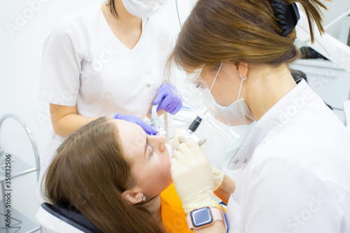 A dentist in his practice or office treats a female patient with an assistant in masks and gloves