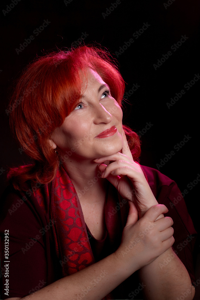 Stylish elderly woman with red hair in vinous dress posing in studio with dark background and red flash and light