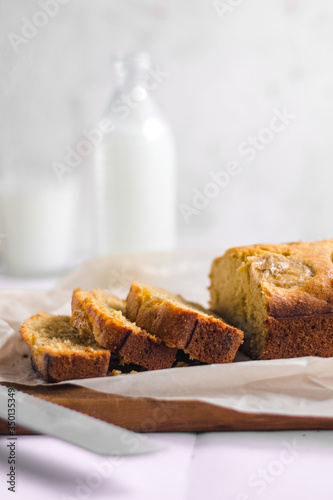 Homemade sliced healthy banana bread ready for serve on breakfast on the white concrete background with craft baking paper and pastry knife, glass of milk. Top view. Close up shoot