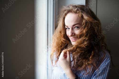 Close-up portrait of young woman standing by window indoors at home.