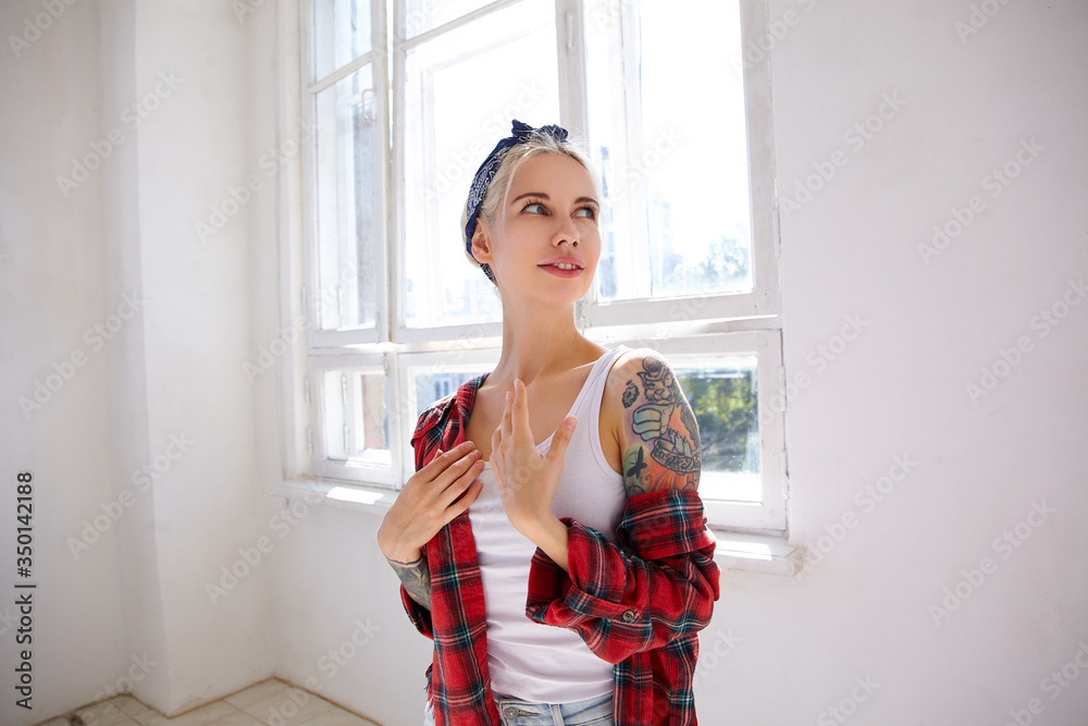 Positive young attractive blonde woman with tattoos dressed in checkered shirt keeping palms on chest while looking gladly aside, posing in front of big window