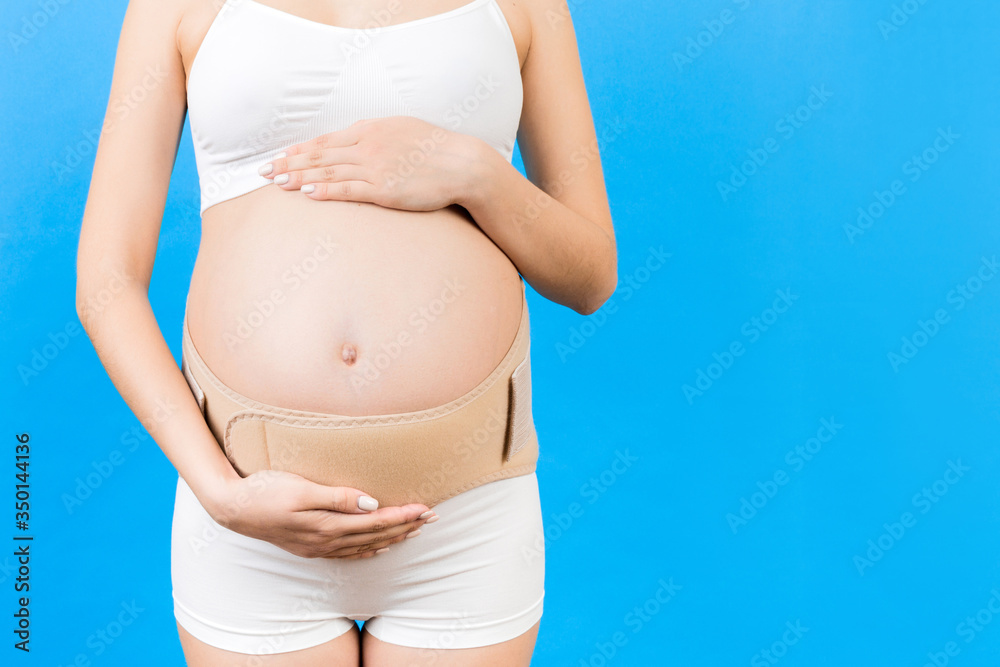 Close up of pregnant woman in underwear dressed in orthopedic corset for reducing pain in the back at blue background with copy space. Orthopedic abdominal support belt concept Close up of pregnant woman in underwear dressed in orthopedic corset for reducing pain in the back at blue background with copy space. Orthopedic abdominal support belt concept