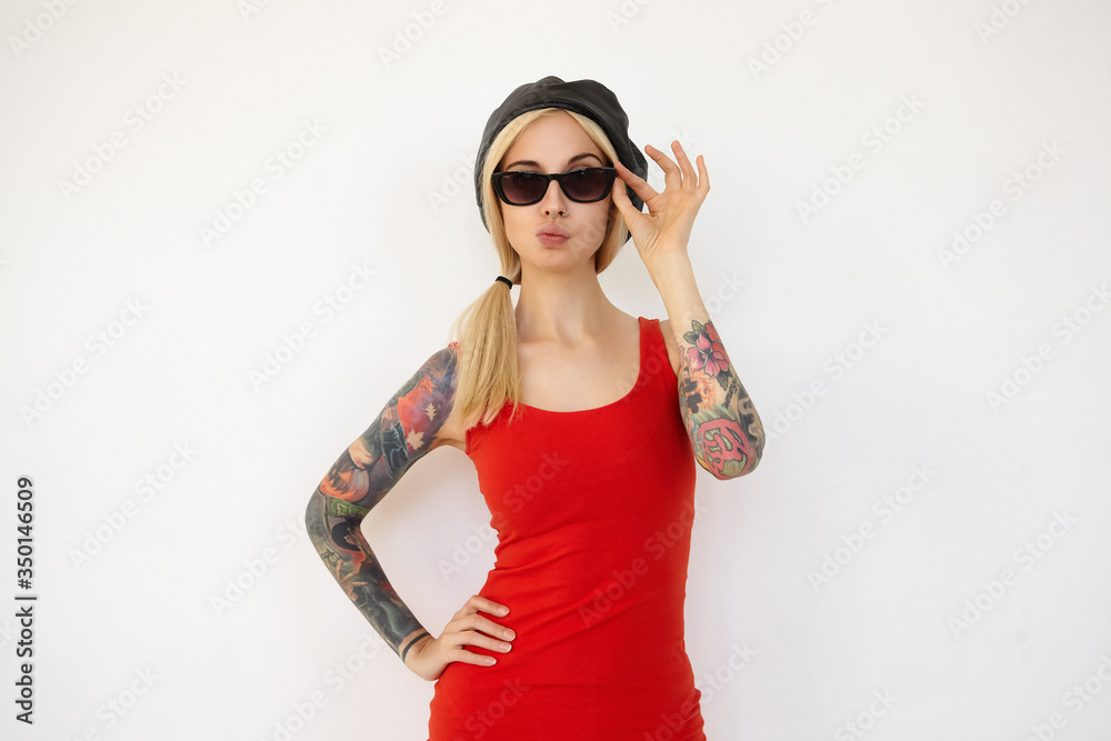 Puzzled young elegant tattooed blonde female in black beret and sunglasses keeping her hand on waist and pursing lips while standing over white background Puzzled young elegant tattooed blonde female in black beret and sunglasses keeping her hand on waist and pursing lips while standing over white background