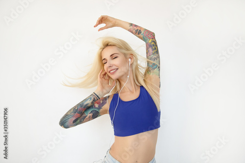 Indoor shot of young beautiful blonde long haired lady with tattoos keeping raising her hands while dancing with music in her earphones  posing over white background