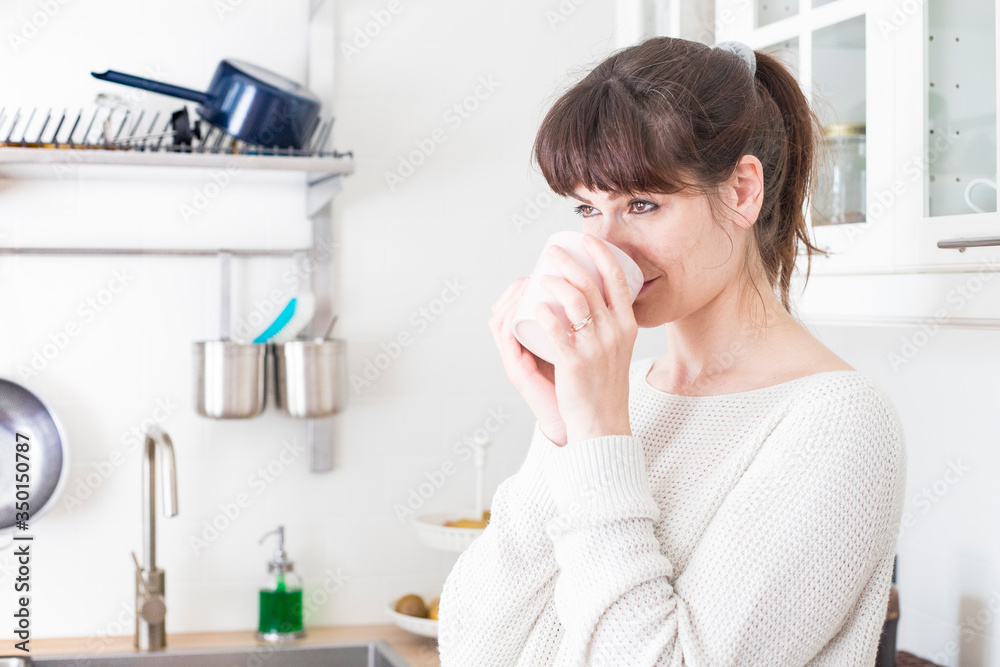 Pretty caucasian woman, with bangs and ponytail, drinking a cup of coffee in the morning in a white, bright lit, kitchen. Profile portrait, smiling. Relaxed and positive atmosphere. Pretty caucasian woman, with bangs and ponytail, drinking a cup of coffee in the morning in a white, bright lit, kitchen. Profile portrait, smiling. Relaxed and positive atmosphere.