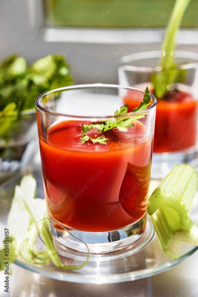 Two glasses of tomato juice with parsley and celery decorations, stand near the window, morning sunlight shines, shallow depth of field, selective focus. Natural drinks concept. Two glasses of tomato juice with parsley and celery decorations, stand near the window, morning sunlight shines, shallow depth of field, selective focus. Natural drinks concept.
