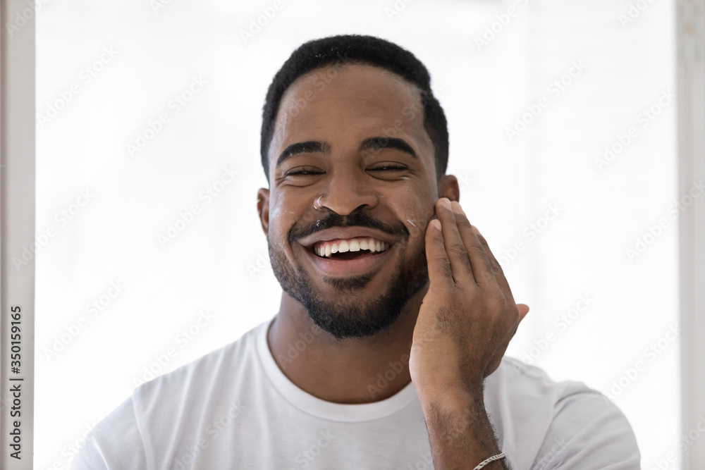 Obraz premium Headshot portrait of smiling young african American man apply nourishing facial cream or balm in bathroom, happy biracial male use moisturizing anti-wrinkle face skin mask, skincare concept