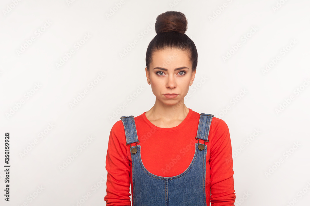 Portrait of upset depressed girl with hair bun in denim overalls looking at camera with serious unhappy face, gloomy sad expression, worrying about problems. studio shot isolated on white background Portrait of upset depressed girl with hair bun in denim overalls looking at camera with serious unhappy face, gloomy sad expression, worrying about problems. studio shot isolated on white background