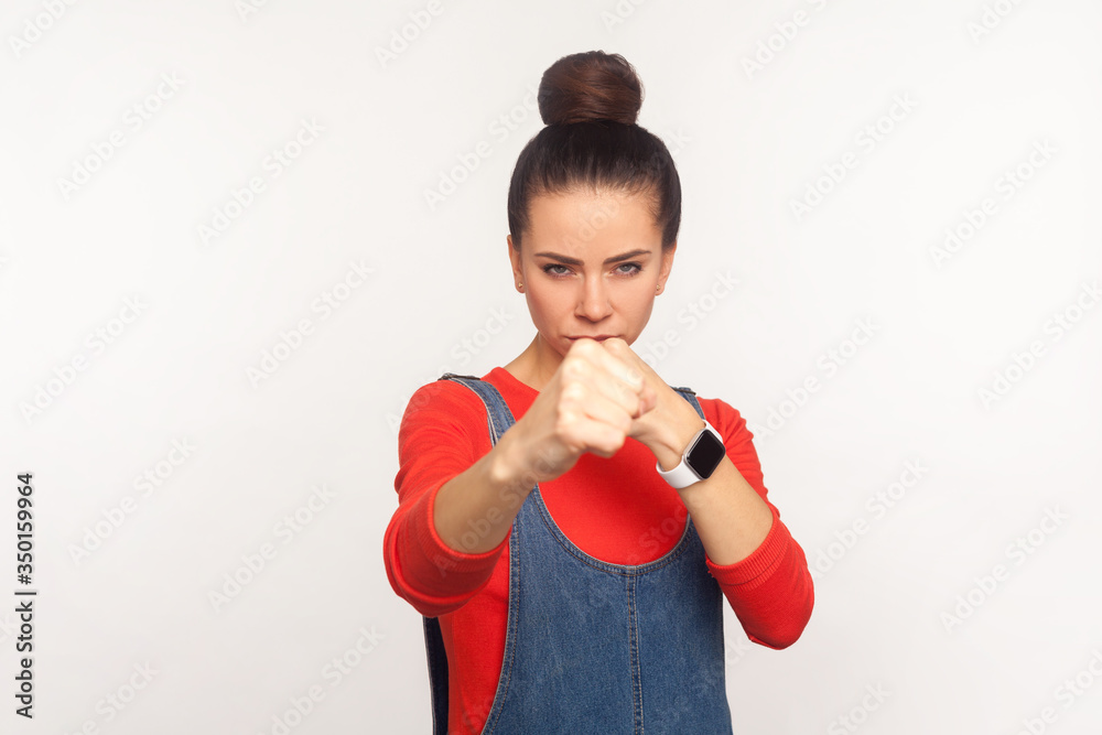 Let's fight! Portrait of determined aggressive confident girl with hair bun in denim overalls boxing with clenched fist, punching in front of camera, struggle and self-defence. studio shot, isolate Let's fight! Portrait of determined aggressive confident girl with hair bun in denim overalls boxing with clenched fist, punching in front of camera, struggle and self-defence. studio shot, isolate