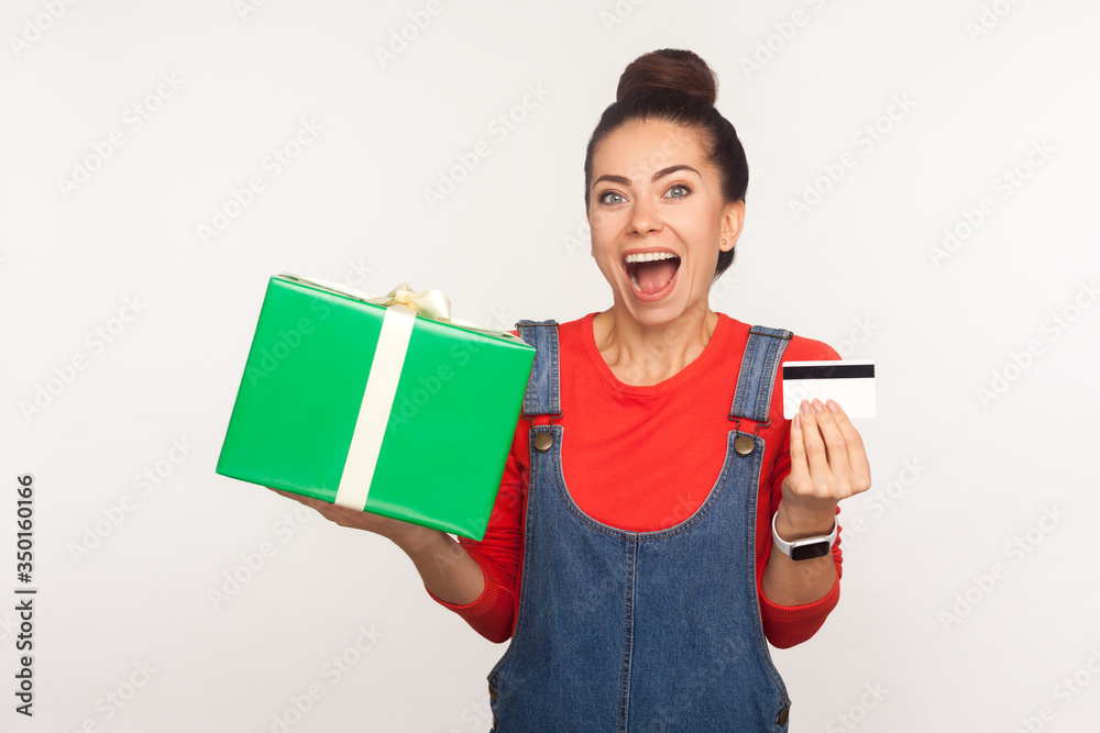 Wow, credit for holiday gift purchase! Portrait of stylish joyful girl with hair bun in denim overalls holding present box and bank card, enjoying shopping with cashback, bonus. studio shot isolated Wow, credit for holiday gift purchase! Portrait of stylish joyful girl with hair bun in denim overalls holding present box and bank card, enjoying shopping with cashback, bonus. studio shot isolated
