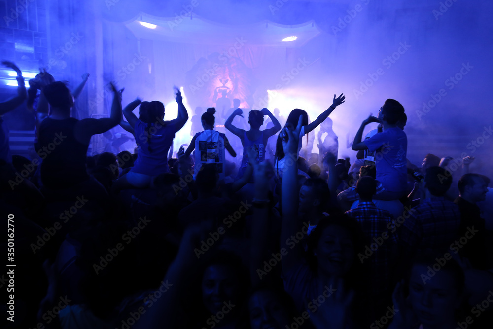 People Raising hands against blue smoky background with light beams during concert and crowd of people dancing.