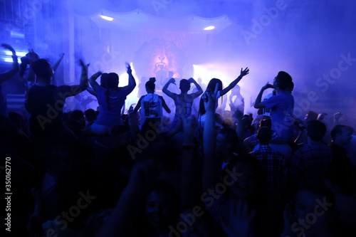 People Raising hands against blue smoky background with light beams during concert and crowd of people dancing.