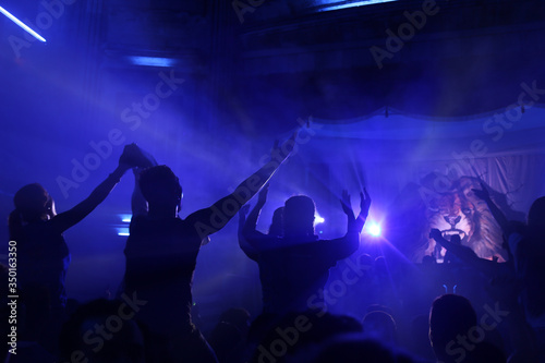 People Raising hands against blue smoky background with light beams during concert and crowd of people dancing.