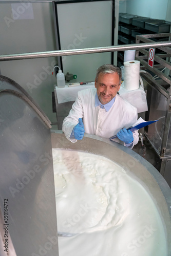 Smiling Dairy Plant Food Engineer Giving Thumbs Up Beside Stainless Steel Milk Pasteurizer Tank