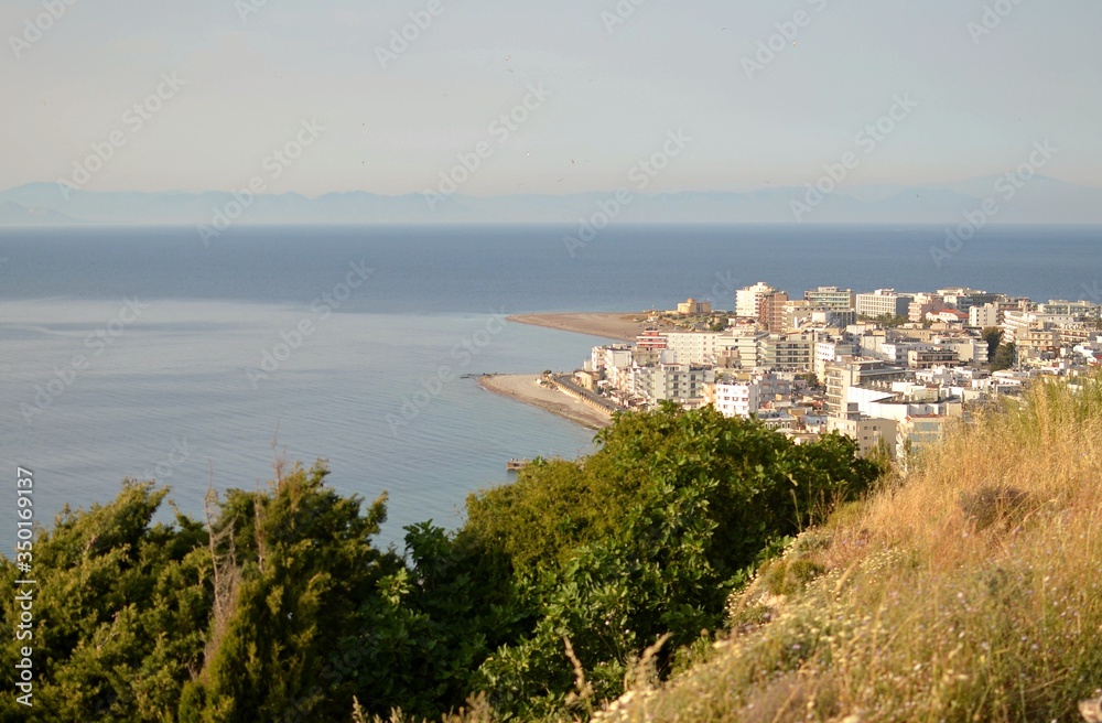 View of the city of Rhodes from the cliff on the island. In the foreground is dried grass with ears of a path. Sea view and blue sky. a view of the multi-storey buildings of the city and the mountains