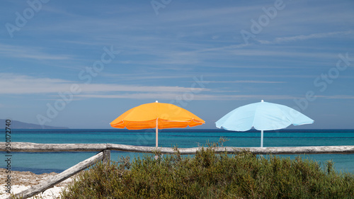 Beach umbrellas blue and orange on the beach behind the wooden fence. Holidays in Sardinia.