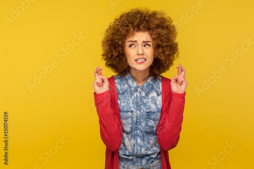 Waiting fortune. Portrait of woman with curly hair crossing fingers for good luck, making wish and hoping for win, clenching teeth in anticipation of success. studio shot isolated on yellow background