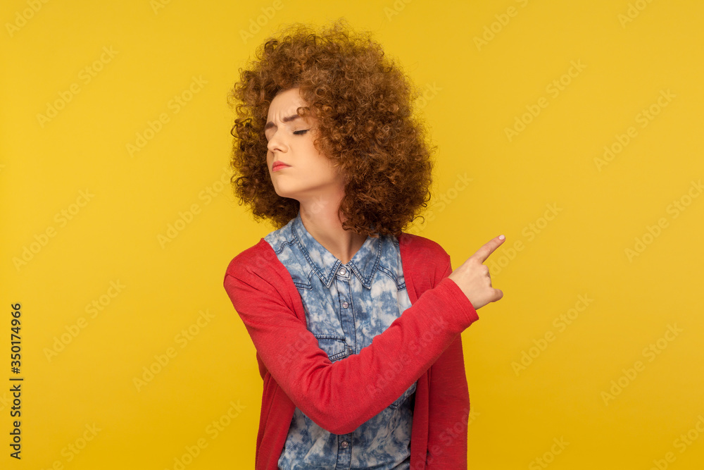 Get out! Portrait of upset vexed woman with curly hair showing exit, demanding to leave her alone, turning away with resentful irritated expression. indoor studio shot isolated on yellow background
