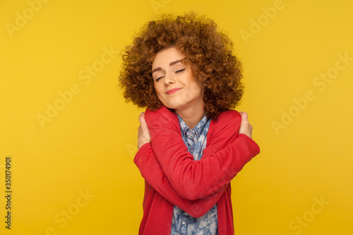 I am the best! Portrait of selfish woman with fluffy curly hair embracing herself and smiling with pleasure expression, self-loving and egoism concept. indoor studio shot isolated on yellow background © khosrork