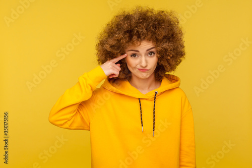 This is crazy! Portrait of curly-haired woman in urban style hoodie making stupid sign with finger near head, gesturing bad mind, dumb insane idea. indoor studio shot isolated on yellow background