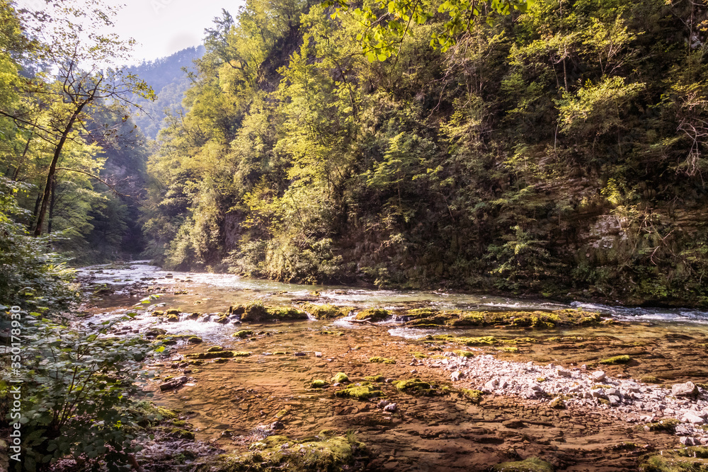 Bluewater mountain river with a small waterfall in the middle with a ray of sun shining through green and yellow trees on right and left sides in Vintgar Gorge, Slovenia during summer