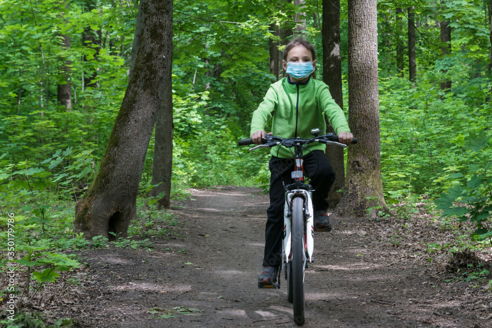 boy in protective medical mask rides a bike in deep green forest, safe new way of sport activities after end of quarantine lockdown,new rules of outdoor sport activities