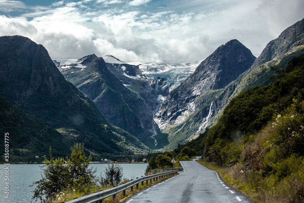 Trip to Norway. Blue ice tongue of Jostedal glacier melts from the giant rocky mountains into the green valley with waterfalls, big lake and asphalt road on background of blue sky & white clouds Trip to Norway. Blue ice tongue of Jostedal glacier melts from the giant rocky mountains into the green valley with waterfalls, big lake and asphalt road on background of blue sky & white clouds