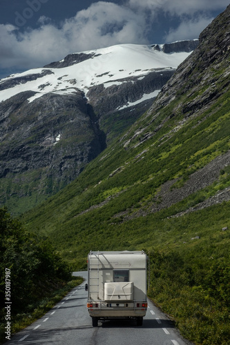 White trailer rides on the alpine road surrounded by rock mountains, snow peaks & green valley on background of blue sky with white clouds in sunny summer day. Vertical wallpaper. Norway