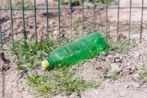 Environmental pollution by household waste. A green plastic bottle lies under iron fences. The concept of environmental issues.