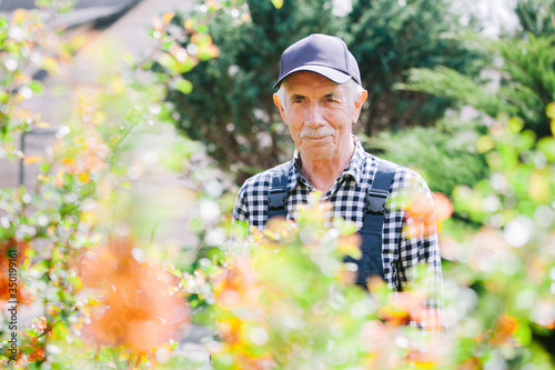 Senior gardener standing in garden. Aged man in overall and baseball cap. Portrait of old farmer