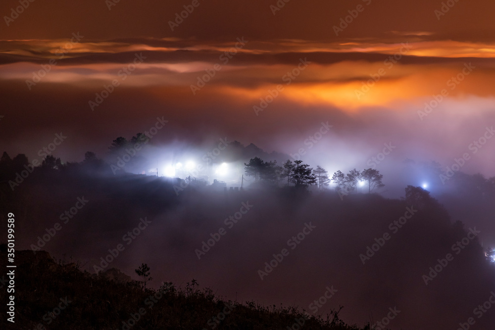 Fototapeta premium Mountains in fog at beautiful night in autumn in Dalat city, Vietnam. Landscape with Langbiang mountain valley, low clouds, forest, colorful sky with stars, city illumination at dusk.
