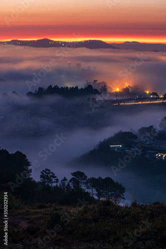 Mountains in fog at beautiful night in autumn in Dalat city, Vietnam. Landscape with Langbiang mountain valley, low clouds, forest, colorful sky with stars, city illumination at dusk. © Nhan