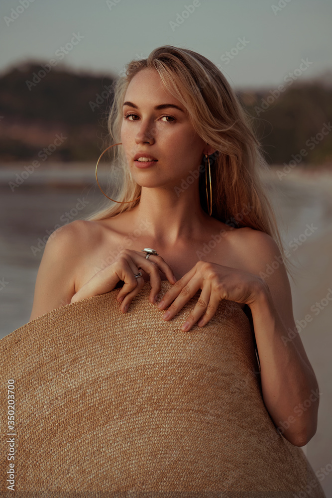 portrait of a blond girl covered herself with a big straw summer hat on the beach and looking at camera