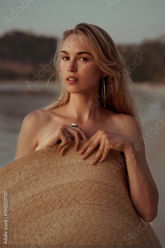 Obraz na płótnie portrait of a blond girl covered herself with a big straw summer hat on the beach and looking at camera w salonie