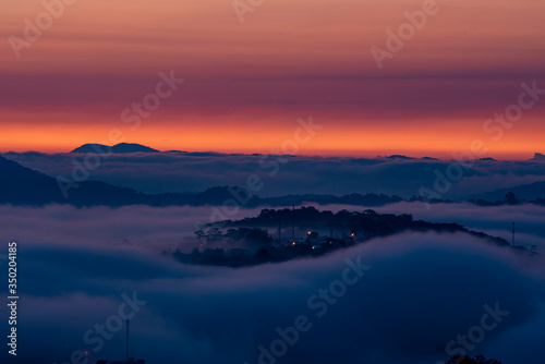 Mountains in fog at beautiful morning in autumn. Landscape with Langbiang mountain valley, low clouds, forest, colorful sky , city illumination at dusk. © Nhan