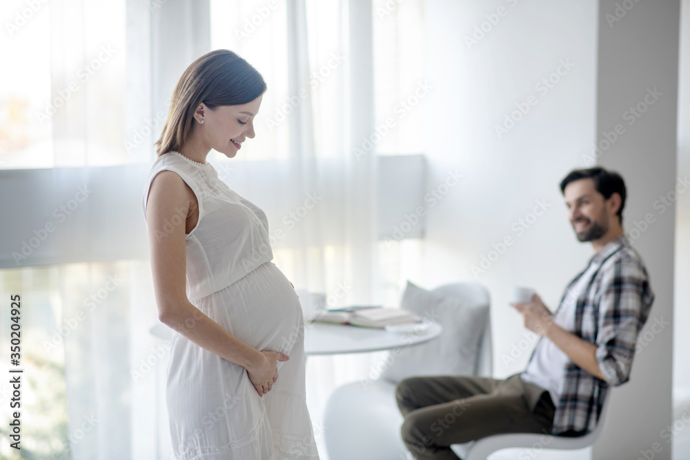 Cute young pregnant woman in a white dress standing in the room