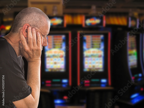 Sad and depressed caucasian man sitting with his hands on his head in front of rows of casino slot machines. Gambling addiction theme image.  Close up portrait. photo